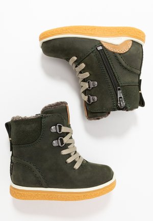 Zielone skórzane botki za kostkę z futrzanym sztucznym wyłożeniem, sznurowanym przodem, zamkiem błyskawicznym oraz gumowymi podeszwami z pomarańczową, teksturowaną środkową częścią podeszwy.