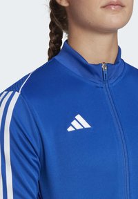 Blue athletic jacket with a high collar, front zipper, and white Adidas logo. Features white stripes on the shoulders and textured fabric.