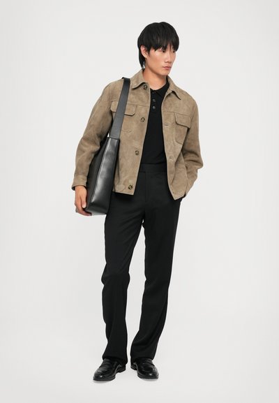 Tan suede jacket with two front pockets, black polo shirt, black dress pants, and black shoes. Holds a textured black shoulder bag.