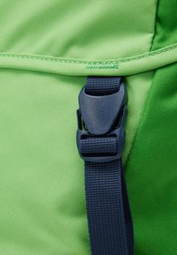 Grön ryggsäck av tyg med en texturerad yta, som har en mörkblå plastspänne och rem för säker stängning. Sömnadsdetaljer synliga.