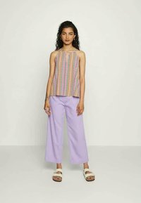 Top de tanque colorido com padrão e decote em forma de chave, combinado com calças largas de tom lilás. Modelo usa sandálias brancas. Fundo minimalista.