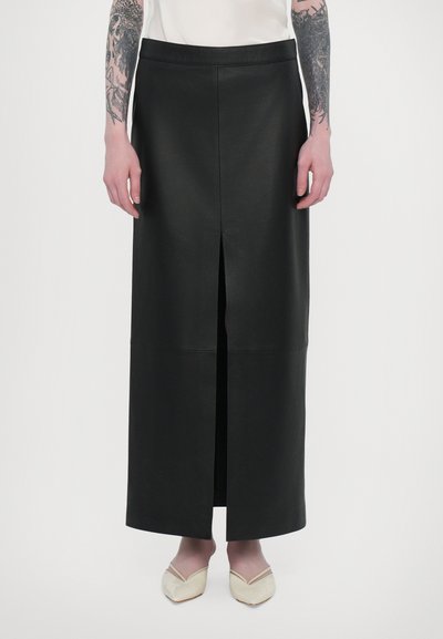 Sort læder maxi-skjørt med en frontslids, der har en tætsiddende talje og en glat overflade. Fodtøjet inkluderer lyse slip-on sko.