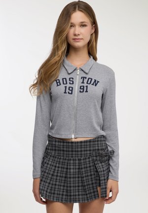 Top corto grigio con zip e texture a coste, con la scritta "BOSTON 1991" stampata in blu navy. Abbinato a una gonna plissettata a quadri neri e grigi con fibbie.