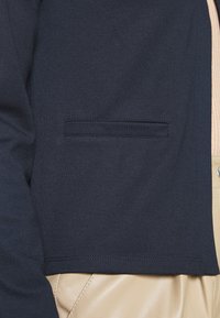 Námořnicky modrá zkrácená bunda z hladkého materiálu, s jednou přední kapsou, na pozadí béžových texturovaných kalhot.