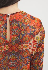 Top rosso a fantasia con scollatura rotonda, dotato di una piccola chiusura a bottone sul retro e un design floreale colorato e testurizzato.