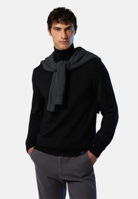 Maglione nero con colletto alto, decorato con una sciarpa grigia drappeggiata sulle spalle. Il modello indossa pantaloni grigi. Tessuto liscio, design aderente.