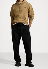 Mand iført tanfarvet Polo Sport hættetrøje, sorte cargobukser med snøre i taljen, sorte sneakers og et armbåndsur, stående med hænderne i lommerne.