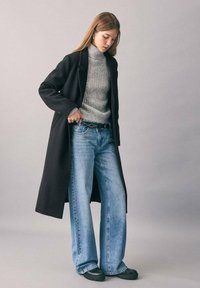 Cappotto lungo nero, maglione grigio a coste con collo alto, jeans larghi in denim blu e scarpe slip-on nere. La cintura con borchie argentate aggiunge un tocco di dettaglio. Sfondo neutro.