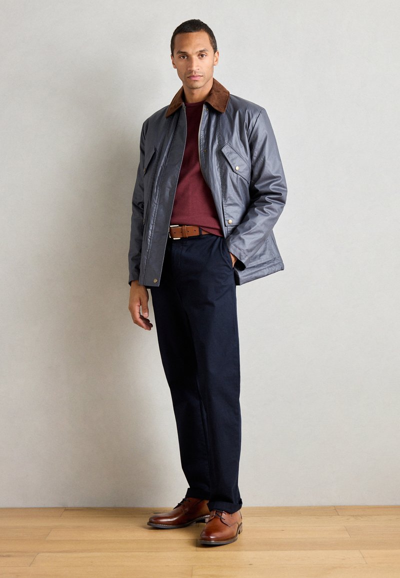 Chaqueta impermeable gris con collar de pana marrón, suéter burdeos, pantalones azul marino y zapatos de cuero marrón, de pie sobre un suelo de madera.