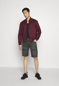 Giacca bomber borgogna sopra una canotta nera, abbinata a un paio di shorts in denim grigio strappato e sneakers nere. Design semplice e moderno.