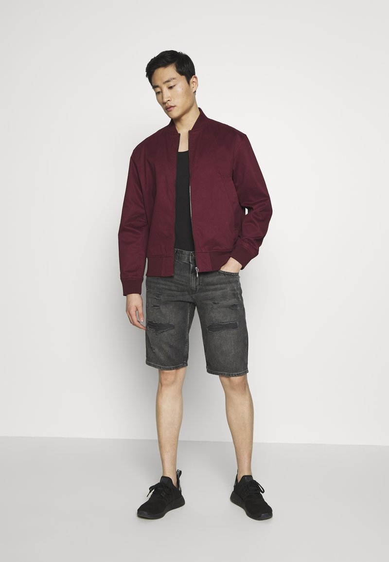 Burgundy bomber-takki mustan hihattoman paidan päällä, yhdistettynä kuluneisiin harmaisiin farkkushortseihin ja mustiin lenkkareihin. Yksinkertainen, moderni muotoilu.