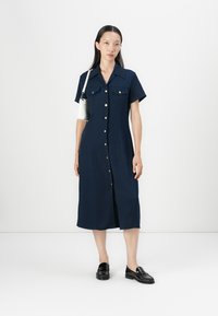 Marineblå knapp-up kjole med korte ermer, to brystlommer og gullknapper. Modellen har på seg svarte loafers og holder en liten hvit veske.