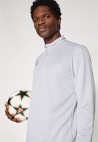 Ανοιχτό γκρι αθλητικό μπλουζάκι με μακριά μανίκια και ψηλό γιακά. Το μοντέλο κρατάει μια λευκή ποδοσφαιρική μπάλα με μαύρα αστέρια και ροζ λεπτομέρειες.