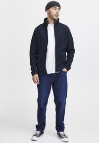 Chaqueta de forro polar azul marino con cuello alto, llevada sobre una camiseta blanca, combinada con vaqueros azules y zapatillas negras, creando un look casual.