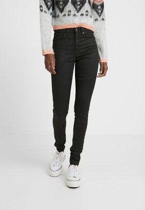 Persoon die strakke zwarte spijkerbroeken, witte platformsneakers en een grijze trui met zwarte en perzikkleurige patronen draagt, staand tegen een effen achtergrond.