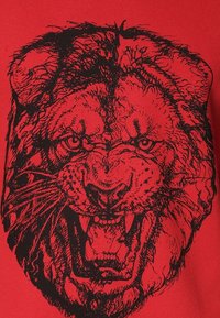 Rotes T-Shirt mit großem schwarzem Löwenmotiv, das detaillierte Gesichtszüge zeigt, einschließlich ausdrucksstarker Augen und ausgeprägter Mähnentextur.