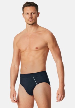 Uomo muscoloso con capelli corti che indossa slip nero con cuciture bianche, in piedi contro uno sfondo chiaro e uniforme.