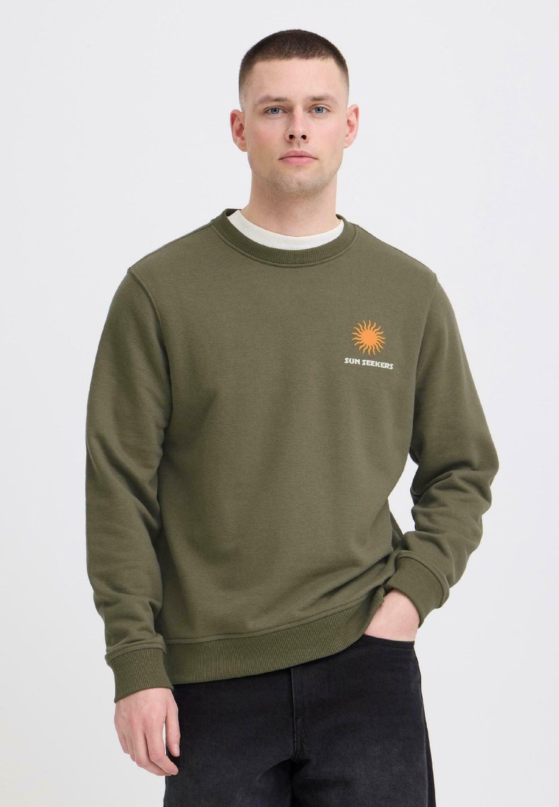 Jeune homme portant un sweat-shirt vert olive avec un soleil orange et le texte "SUN SEEKERS", associé à un pantalon noir, debout devant un fond blanc.