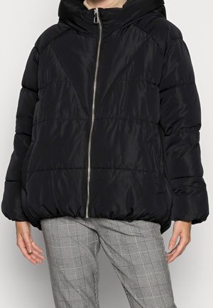 Chaqueta puffer negra con cuello alto, cierre de cremallera y dobladillo fruncido; el diseño acolchado añade textura. Combinada con pantalones de cuadros.