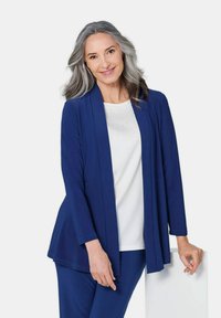 Langer navyfarbener Cardigan aus einem weichen Stoff, mit offenem Frontbereich und langen Ärmeln, über einem weißen Oberteil mit einem subtilen Perlenmuster getragen.