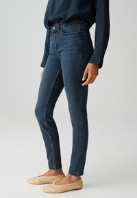 Person trägt dunkelblaue Skinny-Jeans, ein marineblaues langärmeliges Shirt und beige flache Schuhe vor einem einfarbigen hellen Hintergrund.