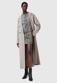 Beiger langer Mantel über einem Kleid mit Schlangenprint, das eine figurbetonte Bodycon-Form, eine glatte Textur und schwarze Stiefeletten mit scharfem Absatz aufweist.