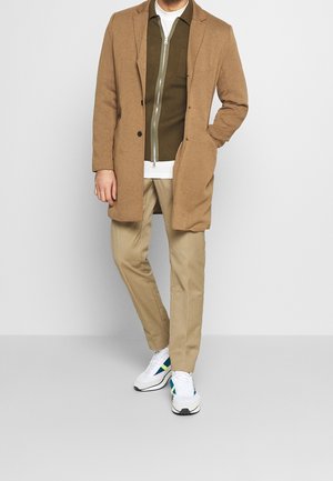 Khaďovně hnědý vlněný kabát, olivově zelená bunda na zip, bílý tričko, khaki kalhoty a bílé sportovní boty s modrými a zelenými akcenty.