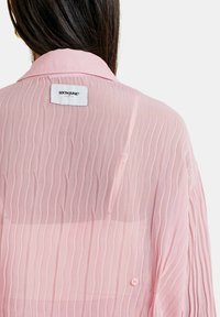 Blusa rosa semitransparente con pliegues verticales. Presenta un collar puntiagudo y un pequeño cierre de botón. Muestra una etiqueta de la marca en la parte posterior.