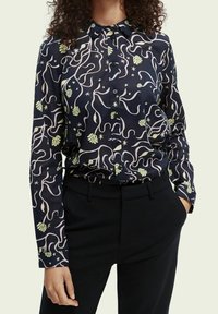 Marineblå skjorte med knapper, prydet med et svunget pastelfarvet og grønt blomstermønster, lange ærmer og traditionel krave, parret med mørke bukser.