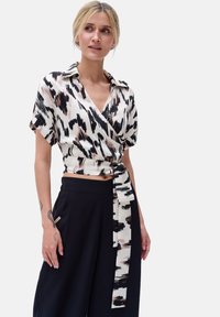 Bluzka w stylu wrap w czarno-białym wzorze z różowymi akcentami, z krótkimi rękawami, kołnierzykiem, marszczonym bokiem z detalem w postaci wiązania, zestawiona z czarnymi spodniami o wysokim stanie.