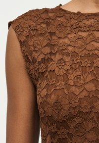 Nahaufnahme eines dunkelhäutigen Arms und einer Schulter, die ein ärmelloses braunes Spitzenkleid mit Blumenmustern tragen.