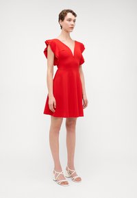 Robe rouge avec un décolleté en V profond, des manches flottantes et une jupe évasée. Tissu lisse et design à hauteur des genoux associé à des talons blancs à lanières.