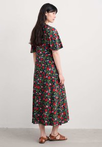 Robe fleurie avec des manches courtes, un corsage cintré et une jupe fluide. Fond vert avec des fleurs rouges et roses, assortie de sandales marron.