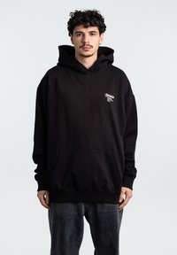 Sudadera de algodón negra con un gran bolsillo frontal, capucha con cordón y un logotipo blanco impreso en el pecho. El modelo lleva pantalones de mezclilla grises.