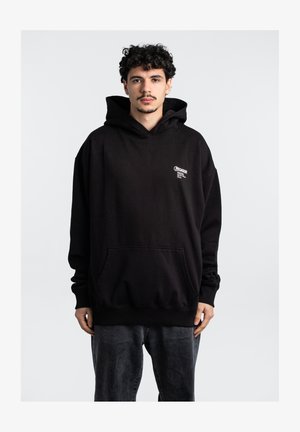 Schwarzer Baumwollhoodie mit großer Fronttasche, Kapuze mit Kordelzug und weißem Logo auf der Brust. Das Model trägt graue Jeans.