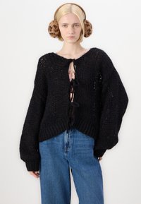 Čierny pletený sveter s voľným strihom, oversized rukávmi a hlbokým V-výstrihom, so zaväzovacími uzávermi. V kombinácii s modrými džínsami.