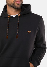 Un primer plano de un hombre que lleva una sudadera con capucha negra con cordones naranjas, destacando un pequeño logo en el pecho. El fondo es blanco liso.