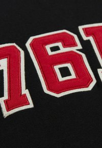 Schwarzer Stoff mit gestickten roten Zahlen "76" und "81", umrandet mit weißen Fäden. Die Textur ist glatt und leicht strukturiert.