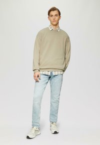 Felpa beige oversize indossata sopra una camicia a quadri chiari, abbinata a jeans azzurri chiari e sneaker crema con accenti grigi.