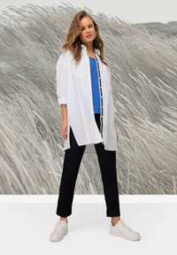 Camicia bianca a maniche lunghe con dettagli gialli sopra una maglietta blu e pantaloni scuri, abbinata a sneakers bianche. Sullo sfondo si vedono erba alta.
