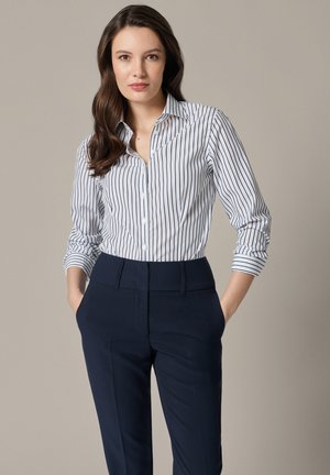 Kobieta z długimi brązowymi włosami, ubrana w białą koszulę z czarnymi pionowymi paskami i granatowe spodnie garniturowe, stoi z rękami w kieszeniach.