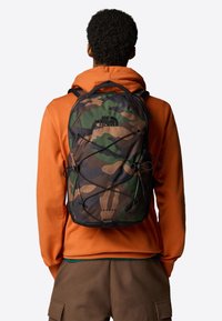 Plecak w kamuflażu o aerodynamicznym designie, z czarnym logo, detalami z elastycznego sznurka oraz wytrzymałym materiałem w odcieniach zieleni i brązu.