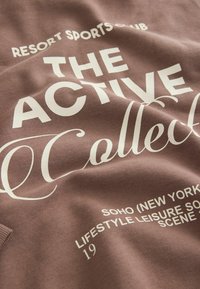 Tessuto marrone con testo stampato bianco, tra cui "LA COLLEZIONE ATTIVA" e "CLUB SPORTIVO RESORT", caratterizzato da una consistenza morbida e un design informale.