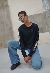 Fekete, hosszú ujjú póló fehér logóval a mellrészen és felirattal az ujjakon, világoskék harangszárú farmernadrággal és fekete, vastag talpú sneaker cipővel párosítva.