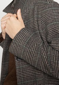Persona che regola il colletto di un cappotto di lana a scacchi marrone e nero, indossando un maglione marrone scuro sotto.