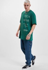 Grøn bomulds T-shirt med "ROCA WEAR ESTABLISHED New York 1999" trykt i hvid. Løs pasform. Modellen bærer blå denimbukser og sorte sneakers.