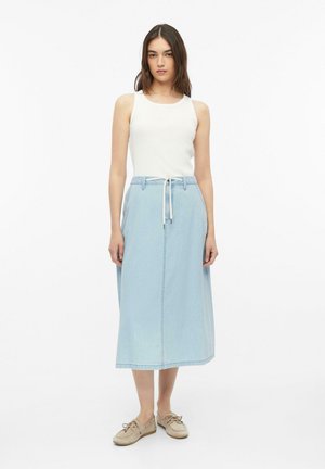 Frau steht, trägt ein weißes ärmelloses Top, einen hellblauen Midi-Denimrock mit Kordelzug und beigen Freizeitschuhen vor weißem Hintergrund.