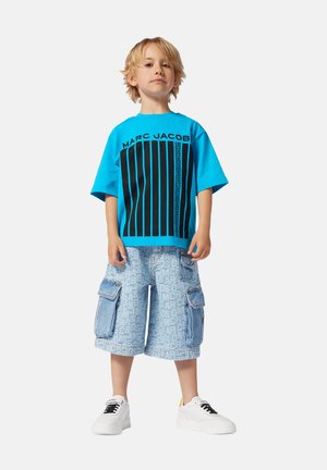 Blauwe T-shirt met zwarte verticale strepen en het logo "MARCBYJACOBS", gecombineerd met licht denim shorts met herhaald tekstpatroon. Witte sneakers.