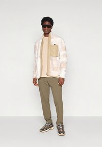 Chaqueta de forro polar con un patrón en crema y beige, bolsillo delantero, combinada con una camisa beige y pantalones verde oliva, completado con zapatillas negras.