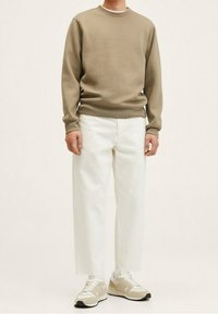 Persona che indossa una felpa beige con scollo rotondo, pantaloni bianchi ampi e corti alla caviglia, e sneakers beige e bianche, in piedi contro uno sfondo chiaro e uniforme.